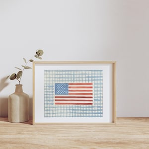 Puede incluir: Pintura en acuarela enmarcada de la bandera estadounidense. La bandera presenta rayas rojas y blancas y un campo azul con estrellas blancas. El fondo es un patrón de cuadros azul claro y blanco. Un jarrón beige con vegetación a la izquierda.