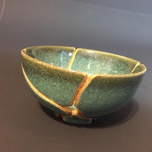 May include: A round ceramic bowl with a mottled green glaze finish. The bowl features visible cracks that have been repaired with a gold-colored material, creating a distinct pattern of lines across the exterior and interior surfaces.