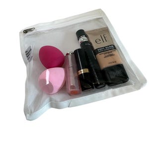 May include: A white mesh zippered pouch with a clear front panel containing makeup products. The products include a pink and a light pink makeup sponge, a clear lip gloss, a black lipstick, and a bottle of elf Soft Glam Satin Foundation.