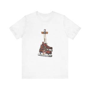 May include: White t-shirt featuring a watercolor illustration of brown ice skates hanging from a wooden sign. The skates have a tag that reads "60". The design is centered on the shirt.