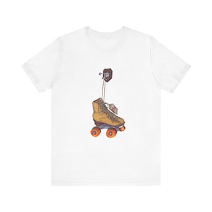 May include: White t-shirt featuring a watercolor illustration of a vintage roller skate hanging from a wall hook. The skate is tan with orange wheels and a tag. The artwork has a retro, nostalgic feel.