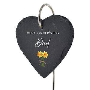 May include: A heart-shaped black slate plaque featuring the white text "HAPPY FATHER'S DAY" and "Dad" above a graphic of two yellow daffodils tied with a brown string. The plaque hangs from a silver metal hook attached to a thin metal rod.