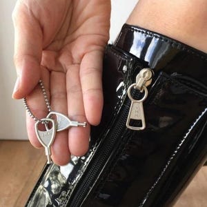 May include: A silver key pendant with two keys on a chain is held near a black patent leather boot with a silver zip. The zip pull has the text "YKK" on it.