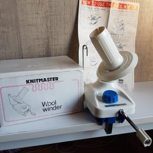 Puede incluir: Enrollador de lana de plástico blanco con una perilla azul y una manivela de metal. El enrollador está sujeto a una mesa con una abrazadera. La caja del enrollador es blanca con letras rosas y dice "Knitmaster Wool Winder".