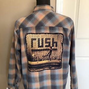 May include: A blue, orange, and brown plaid flannel shirt with a Rush album cover patch on the back. The patch features a person standing in front of a wall of dice.