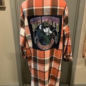 May include: Orange, brown, and white plaid flannel shirt with a Tom Petty and the Heartbreakers patch on the back. The patch features an image of Tom Petty with the band's name. The shirt has long sleeves and a button-down front.