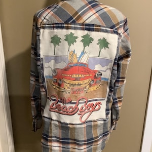 May include: A blue, brown, and white plaid flannel shirt with a vintage The Beach Boys graphic on the back. The graphic features a red car with surfboards on top, palm trees, and the text "The Beach Boys".