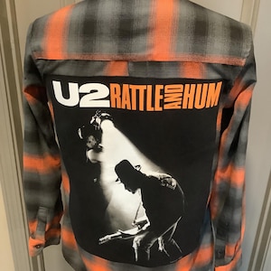 May include: Orange, black, and gray plaid button-down shirt with a large black graphic on the back. The graphic features the text "U2 RATTLE AND HUM" and a black and white image of a musician playing a guitar.