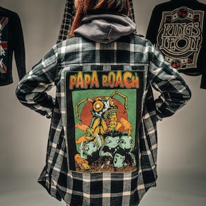 May include: Black and white plaid flannel shirt with a "PAPA ROACH" patch featuring a cartoon insect and figures. Other jackets with graphic designs hang in the background. The shirt has long sleeves and a button-down front.