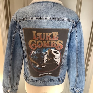 May include: Light blue denim jacket with a cream-colored sherpa collar. The back features a large patch with the text "LUKE COMBS" in a distressed font, along with an image and the text "ASHEVILLE, NC".