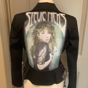 May include: Black blazer featuring a portrait of Stevie Nicks on the back. The image is set against a teal and white background, with the singer's name above. The blazer has a peplum waist and lace-up detailing on the sides.