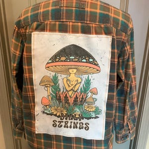 May include: A long-sleeved, button-down flannel shirt in a plaid pattern of green, orange, and brown. The back features a white patch with a colorful illustration of mushrooms and the text "Billy Strings".
