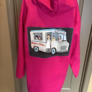 May include: A bright pink hoodie dress featuring a cartoon image of the Bob's Burgers food truck on the back. The food truck graphic is rectangular and has a dark gray border. The hoodie has a hood and long sleeves.