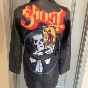 May include: Black leather jacket with the word "Ghost" in red and yellow lettering on the back. A circular patch features a black and white illustration of a figure and a tarot card. The jacket has a belt and is displayed on a mannequin.