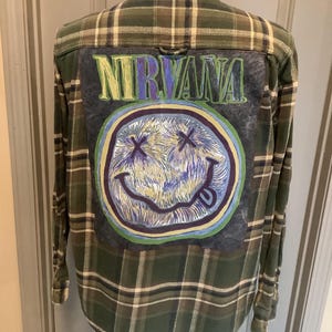 May include: Green and white plaid flannel shirt with a Nirvana graphic on the back. The graphic features the band's name in green and blue, along with a smiley face design with crossed-out eyes and a tongue sticking out.