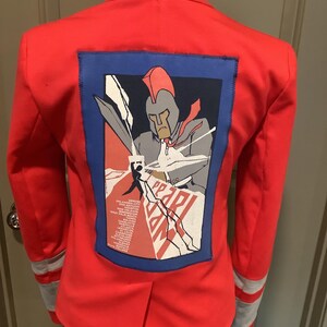 May include: A bright red blazer featuring a large rectangular patch on the back. The patch displays a graphic of a warrior in a helmet holding a sword, with the text PEARL and a list of cities printed below. The sleeves have two horizontal grey stripes.