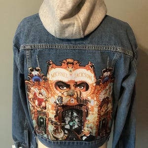 May include: A blue denim jacket with a light gray jersey hood. The back features a large, colorful graphic print of the Michael Jackson Dangerous album cover, including the text Michael Jackson and Dangerous, depicting various figures and eyes.