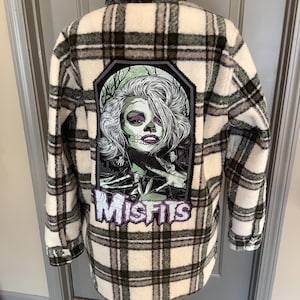 May include: A plaid flannel shirt in cream, black, and gray, featuring a large graphic on the back. The graphic is a stylized illustration of a woman with skeleton makeup, the word "Misfits" below. The shirt is displayed on a mannequin.