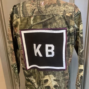 May include: Camouflage button-down shirt with a black square patch featuring white letters "KB". The patch is outlined with a white border and a pink stitched edge. The shirt has a brown and green camouflage pattern.