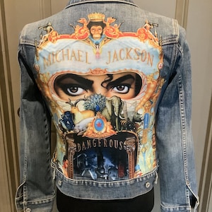 May include: A light-wash denim jacket featuring a colorful graphic on the back. The graphic includes the words "MICHAEL JACKSON" and "DANGEROUS", along with an image of Michael Jackson's eyes and other illustrations. The jacket has a classic button-up design.
