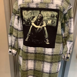 May include: A long-sleeved, button-down shirt featuring a green, white, and black plaid pattern. The back displays a large, rectangular black patch with a graphic print of a person wearing sunglasses, sitting down, with the text HELLO SQUAD.