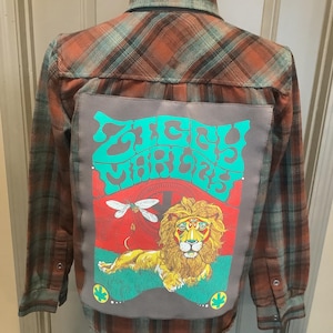May include: Plaid flannel shirt in shades of orange, brown, and teal. The back features a gray patch with the words "Sticky Vibes" in teal, along with a lion illustration, a dragonfly, and other graphics. The shirt has long sleeves and button closures.