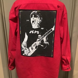 May include: A red button-down shirt with a black and white graphic on the back. The graphic features a musician playing a guitar with the word "DEVO" printed on the shirt. The shirt has long sleeves and buttoned cuffs.