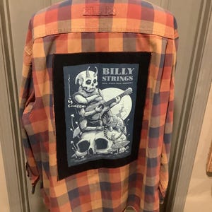 May include: A long-sleeved, button-down flannel shirt in a plaid pattern of orange, red, and blue. A large, rectangular patch on the back features a skull playing a guitar with the text "BILLY STRINGS" in white.