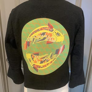 May include: Black jacket with a textured fabric and three-quarter sleeves. The back features a circular design with two yellow koi fish, accented with orange, red, and white details, set against a green and orange wave pattern.