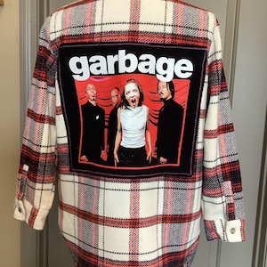 May include: A long-sleeved, button-down flannel shirt in a red, black, and white plaid pattern. The back features a black square patch with the band "Garbage" and a band photo. The shirt has buttoned cuffs.