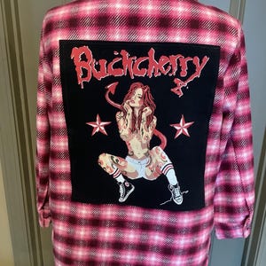 May include: Pink and black plaid flannel shirt with a large black patch on the back. The patch features the band name "Buckcherry" in red script above a tattoo-style illustration of a woman with devil horns and stars.