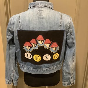 May include: Light blue denim jacket with a black patch featuring a graphic of five figures wearing red hats. The word "DEVO" is spelled out in yellow letters below the graphic. The jacket is displayed on a mannequin.