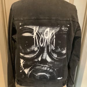May include: A charcoal gray denim jacket featuring a large rectangular back patch with a black and white graphic of a gas mask and the band name Korn printed in white text across the top of the mask design.