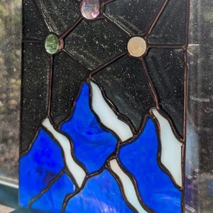 May include: Stained glass panel depicting a mountain range with blue peaks and white snow, set against a black starry sky. The panel includes three colourful glass orbs and is framed with dark brown lines.
