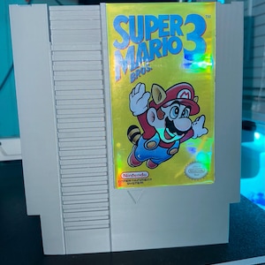May include: A gray Nintendo Entertainment System cartridge for Super Mario Bros. 3. The label features a yellow background with the game title and a cartoon image of Mario in a raccoon suit. The Nintendo logo is visible.