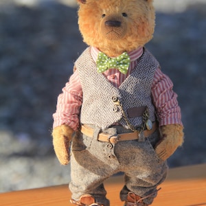 May include: A golden-brown plush teddy bear wearing a pink and white striped shirt, a green polka-dot bow tie, a grey herringbone vest with a gold chain, brown trousers with a tan belt, and brown leather lace-up shoes with visible stitching.