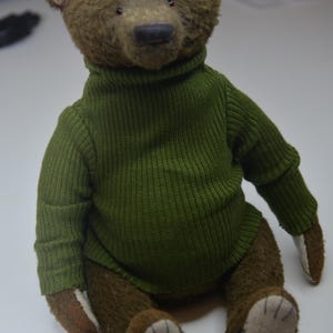 A brown teddy bear wearing a green sweater is sitting on a white surface.
