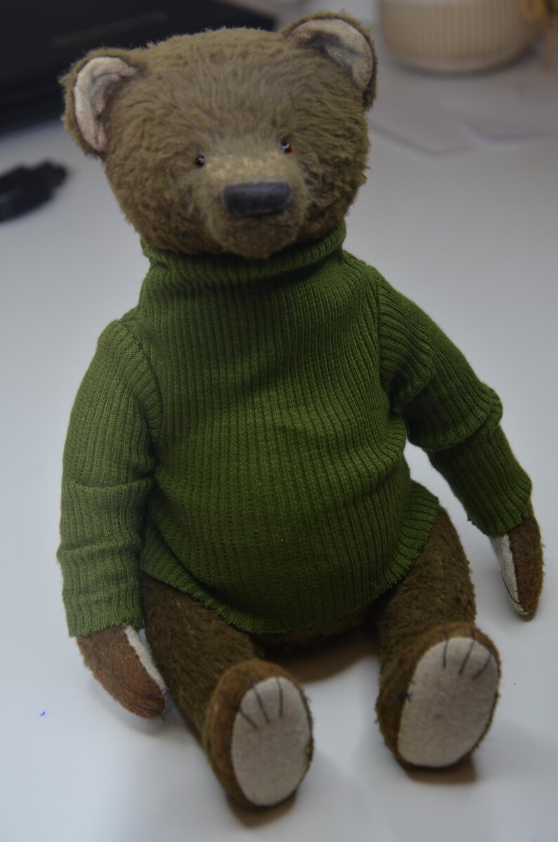 A brown teddy bear wearing a green sweater is sitting on a white surface.