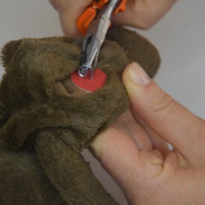 A person's hand is using a pair of scissors to cut the ear of a brown stuffed animal.