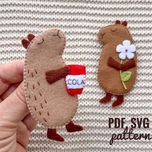 May include: Two felt animal patterns for sewing, one holding a red cup with the word "COLA" on it, the other holding a white flower. The text "PDF SVG pattern" is below the animals.