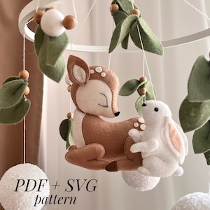 Può includere: Un mobile in legno bianco con un cervo marrone, un coniglio bianco e foglie di feltro verde. Il mobile ha anche pompon bianchi e perline di legno marrone. Il testo "PDF + SVG pattern" è visibile nella parte inferiore dell'immagine.