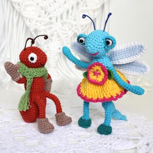 May include: Two crocheted figures: a red ant wearing a light green scarf and brown boots, and a blue insect with white wings, a yellow skirt with a pink border, and a large yellow and pink flower detail on the front, both with dark antennae.
