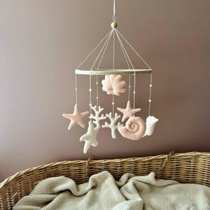 May include: A pink and white felt baby mobile with a wooden ring, featuring a seashell, starfish, coral, and a snail. The mobile is hanging above a wicker basket.