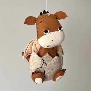 May include: A brown felt dragon with a beige felt egg, hatching from the egg. The dragon has a beige felt face with black eyes and a small brown felt nose. The dragon has beige felt wings and brown felt feet.
