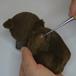 A person's hand is carefully removing a button from a stuffed animal, using a small tool to make the task easier.