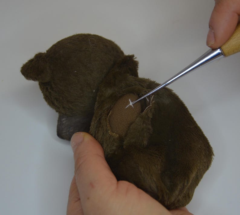 A person's hand is carefully removing a button from a stuffed animal, using a small tool to make the task easier.