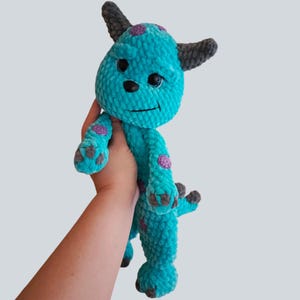 May include: A turquoise plush monster toy with a soft, textured yarn finish. The creature features dark gray horns, purple spots on its body and limbs, a black nose, and dark gray claws on its feet and hands. It has a stitched mouth and eyes.