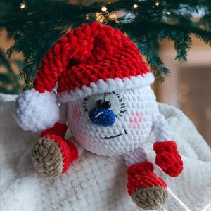 May include: A round, white crocheted plush toy wearing a red and white Santa hat with a pom-pom. The toy features a blue nose, black bead eyes, stitched facial details, pink cheeks, and red mittens and boots with tan-coloured soles.