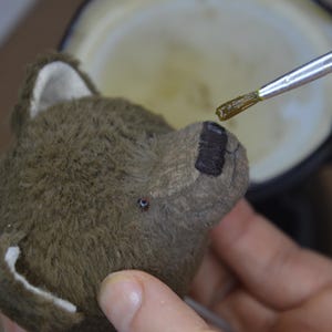 A person is painting a brown teddy bear with a brush, focusing on the bear's face.