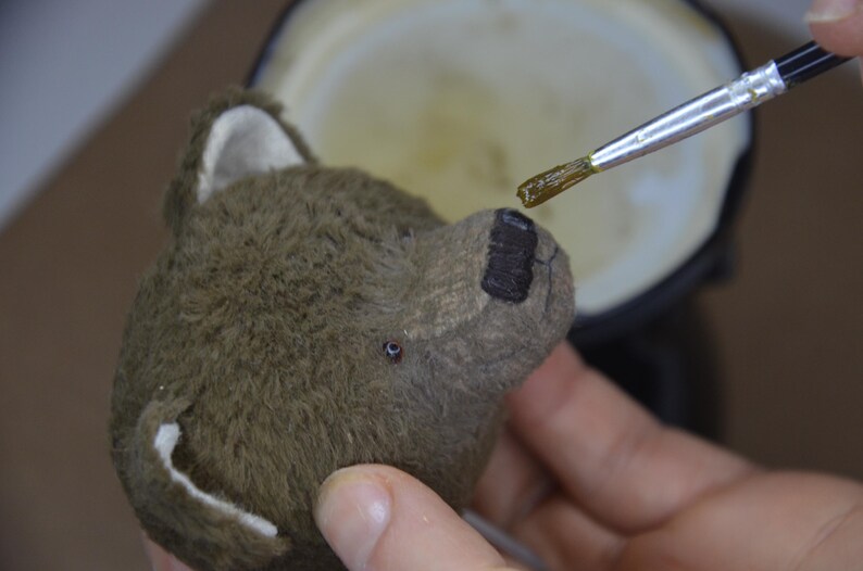 A person is painting a brown teddy bear with a brush, focusing on the bear's face.
